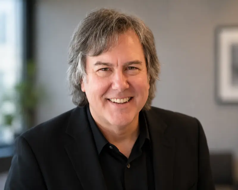 Smiling middle-aged man with gray hair in a black suit, posing for a professional headshot indoors.